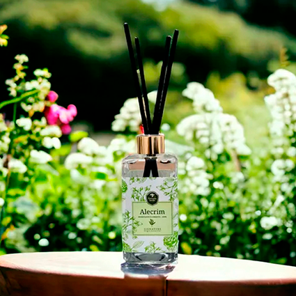 Aromatizador de alecrim em vidro exposto em cima de uma mesa, com um jardimde flores ao fundo.